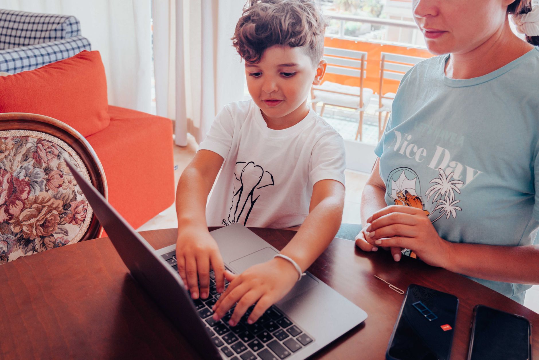 A parent working on a laptop while their child uses a tablet in a home environment.