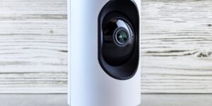 A sleek, modern smart camera placed on a home shelf in a bright, sunlit room.