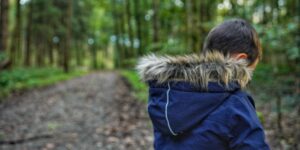 A child standing at the intersection of a forest path and a school building, representing the transition between nature-based learning and formal primary school.